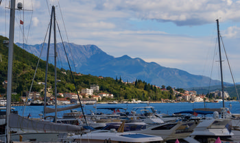This landscape photograph captures the small harbour of Herceg Novi in Montenegro during a bright summer afternoon. The scene prominently features several boats and yachts moored in the harbor, with the picturesque town of Herceg Novi stretching along the coastline of Boka Bay in the background. Nestled between lush green hillsides and beautifully framed by the dramatic Orjen mountain range, which is a notable landmark in the region, the townscape showcases a mix of traditional Mediterranean architecture and modern buildings. The clear blue waters of Boka Bay add to the tranquil atmosphere, while the partly cloudy sky and vibrant greenery reinforce the summery feel of Montenegro’s Adriatic coast.